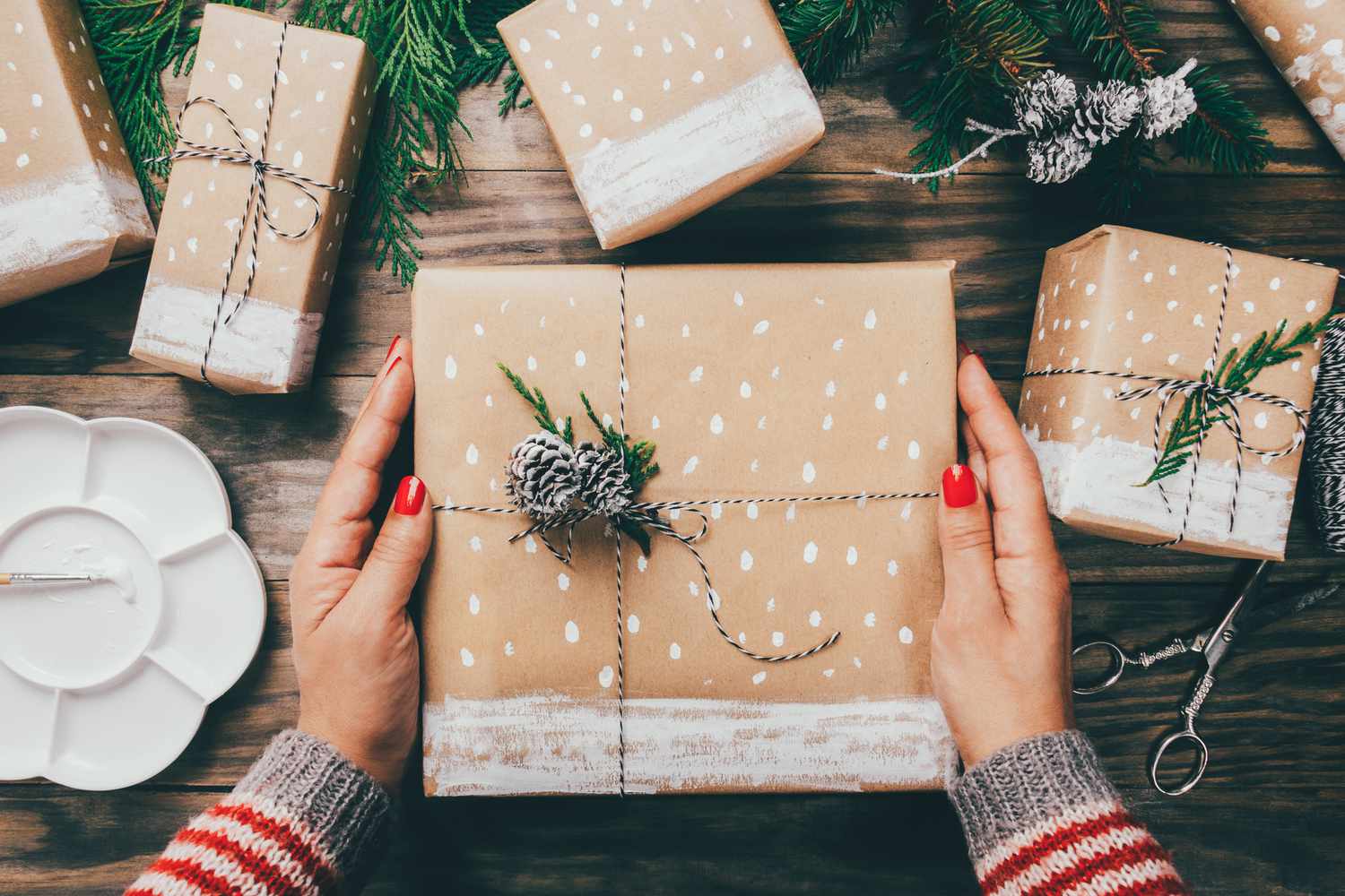 Eine Draufsicht auf eine Frau mit rotem Nagellack, die ein in braunes Papier mit weißen Akzenten eingewickeltes Geschenk hält.