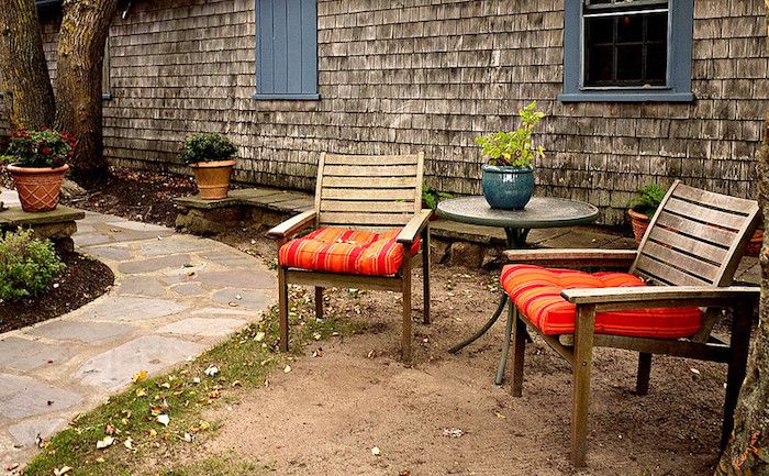 Sillas de madera con cojines naranja brillante junto a un camino de piedra con plantas en macetas sobre mesas y bancos