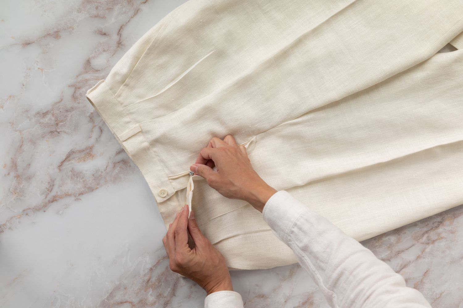 Pantalones color crema con pliegues colocados sobre superficie jaspeada blanca siendo subidos con cremallera 