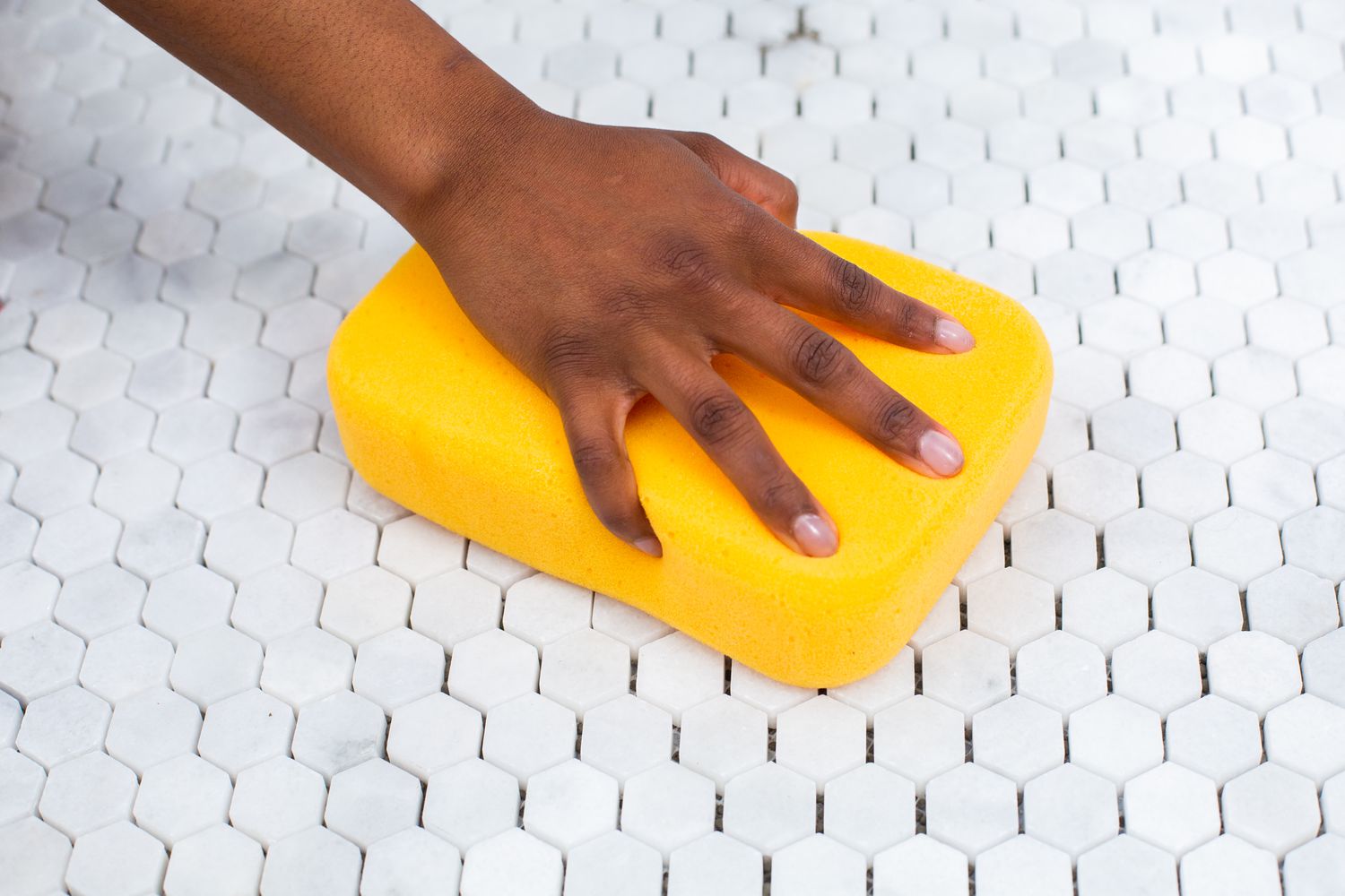 Esponja amarilla con líquido quita bruma de lechada pasando por azulejo de mosaico blanco