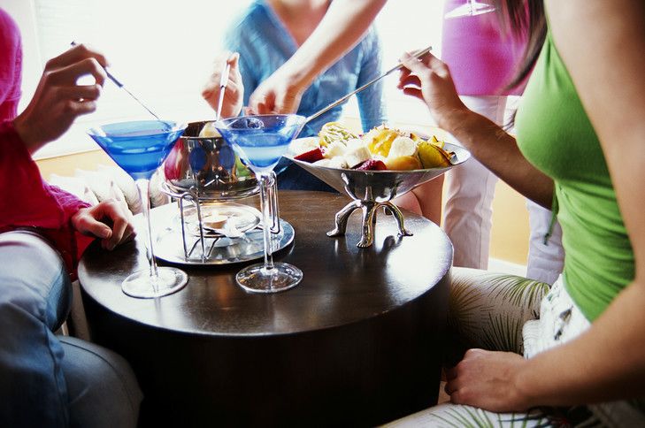 Três pessoas com camisas coloridas reunidas em torno de uma mesa redonda de madeira com coquetéis e fondue