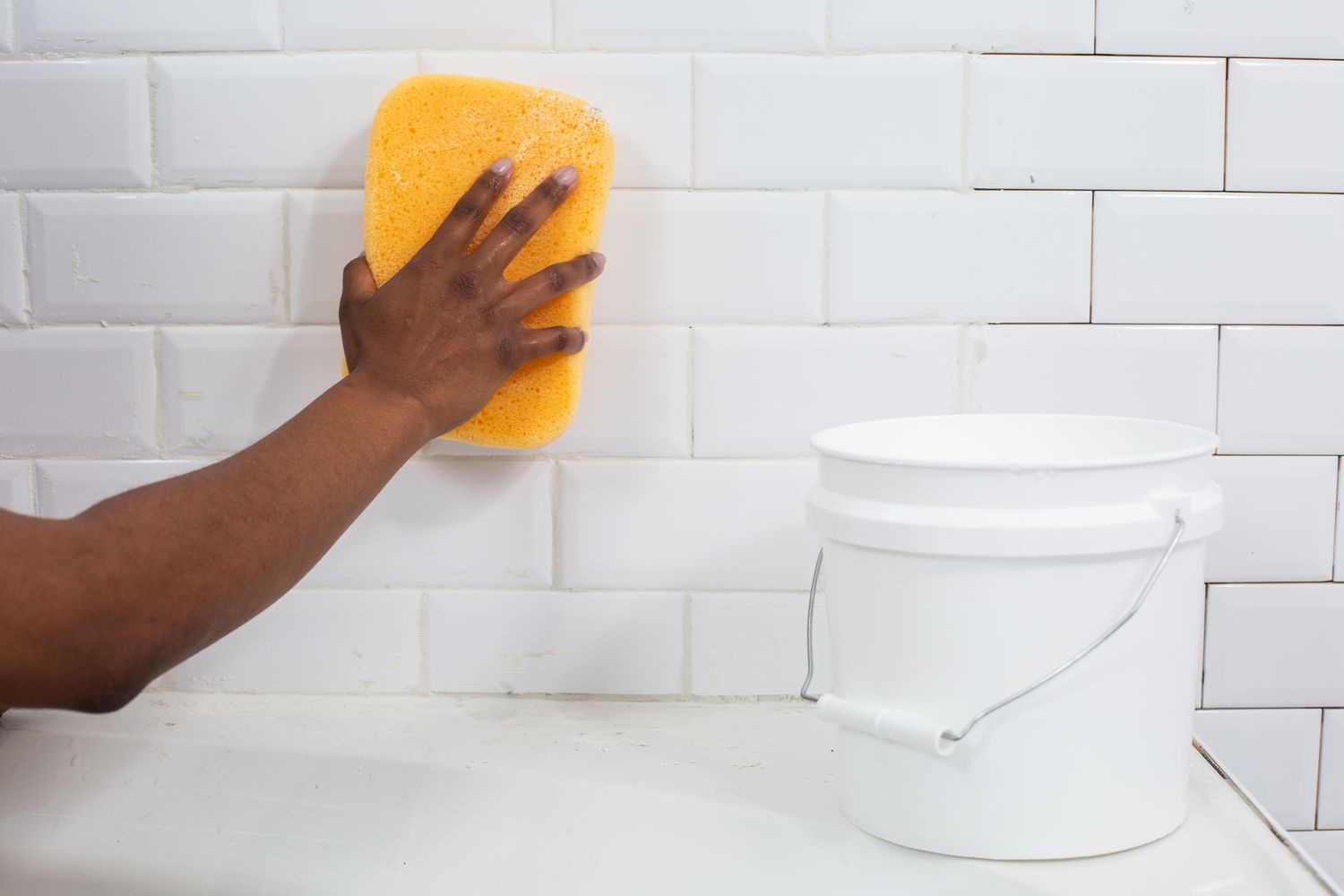 Yellow sponge wiping across white ceramic tile next to white bucket with hot water