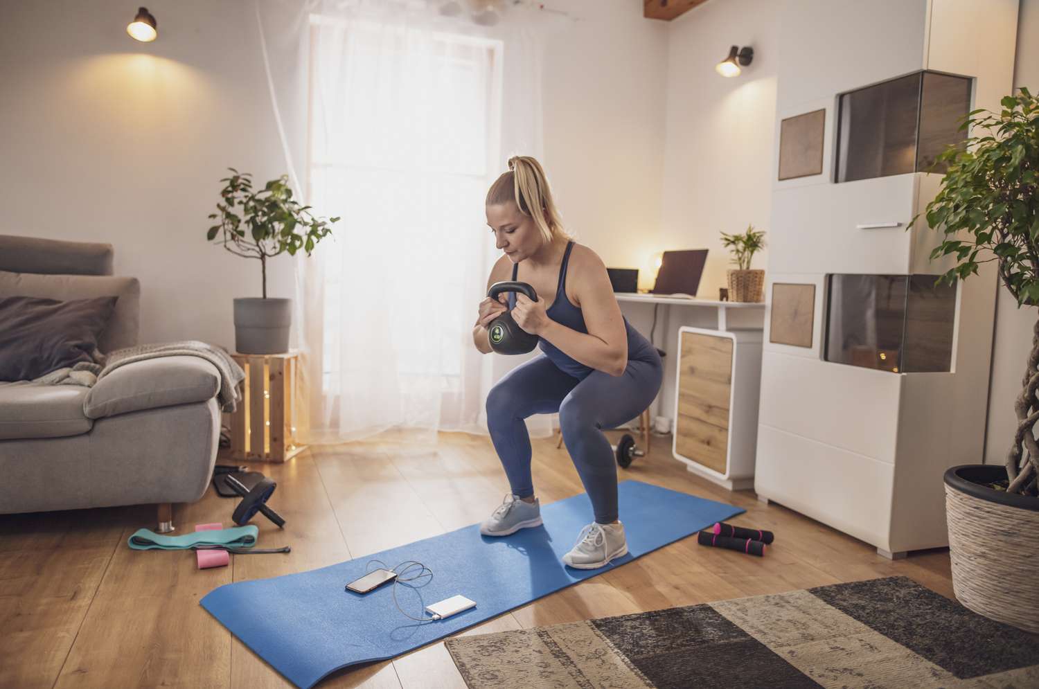 Junge Frau hockt mit Kettlebell auf Yogamatte im Wohnzimmer