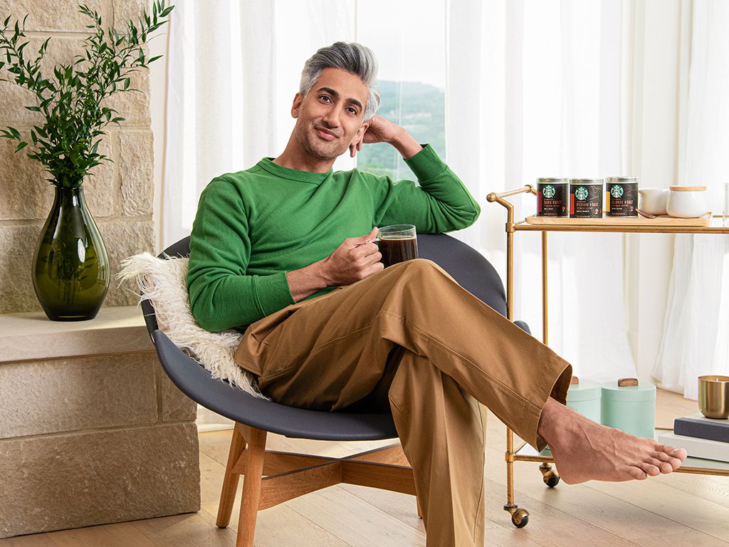 Tan France posiert in seinem Wohnzimmer neben einem Barwagen mit Starbucks-Kaffee. Er hält einen durchsichtigen Kaffeebecher in der Hand und trägt einen grünen Pullover und eine Kamelhose