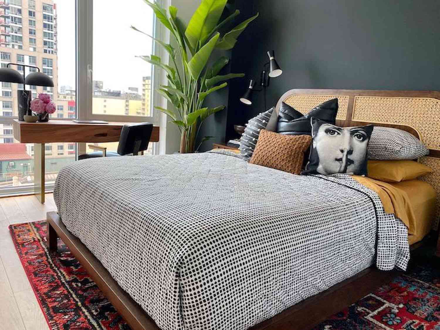 dormitorio moderno con cabecero de mimbre, edredón de lunares blancos y negros, alfombra con estampado rojo, planta grande en una esquina
