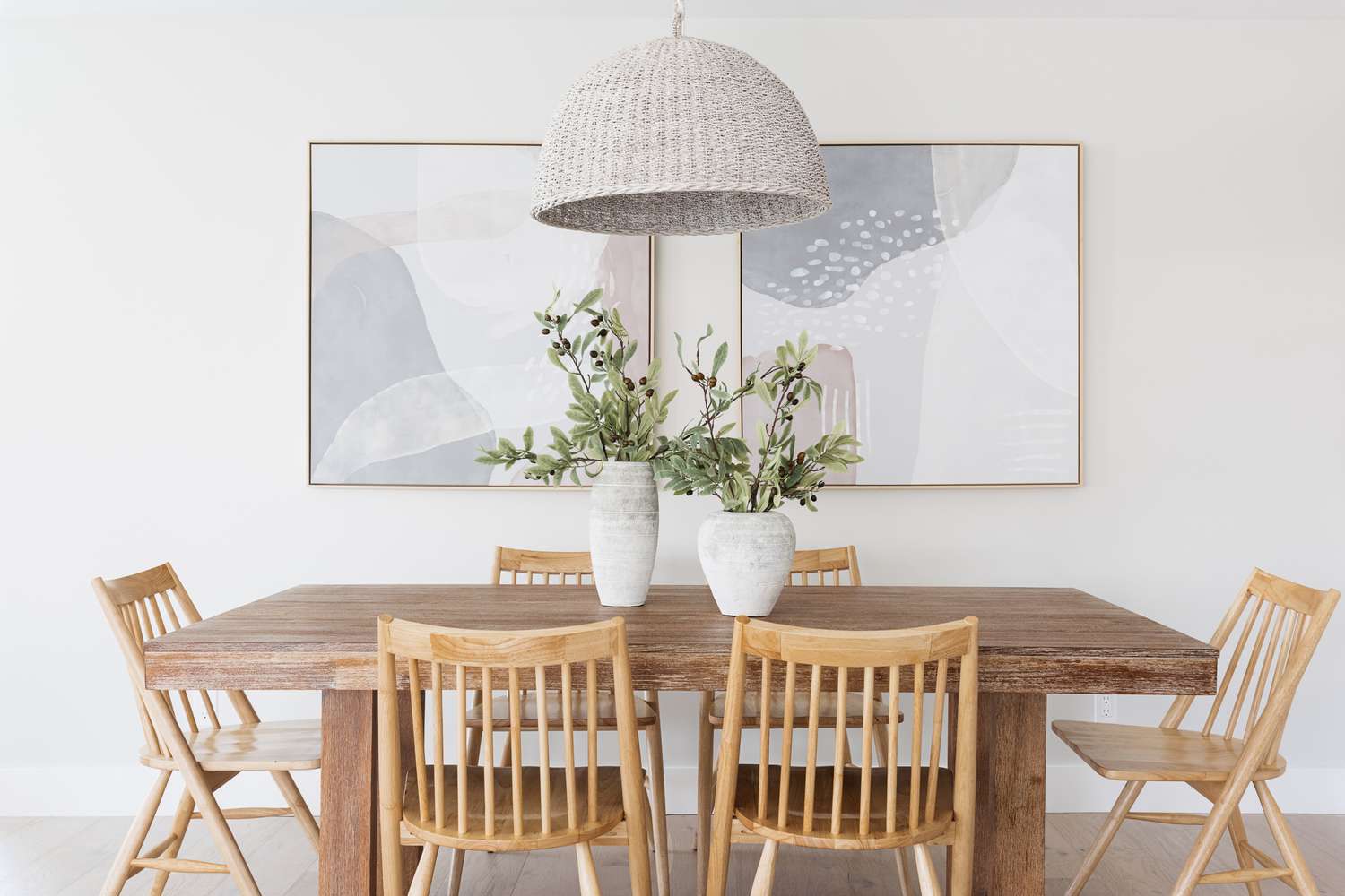 Esszimmer mit Holztisch und Stühlen mit weißen Vasen mit Pflanzen in der Mitte vor weißen Wänden und Gemälden