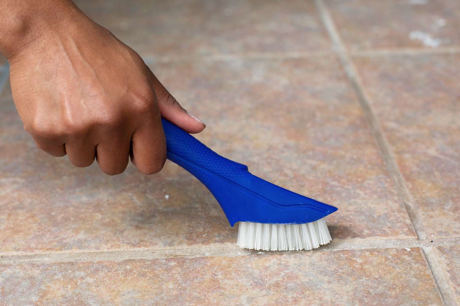 Blue hand-held brush cleaning grout between tile floor