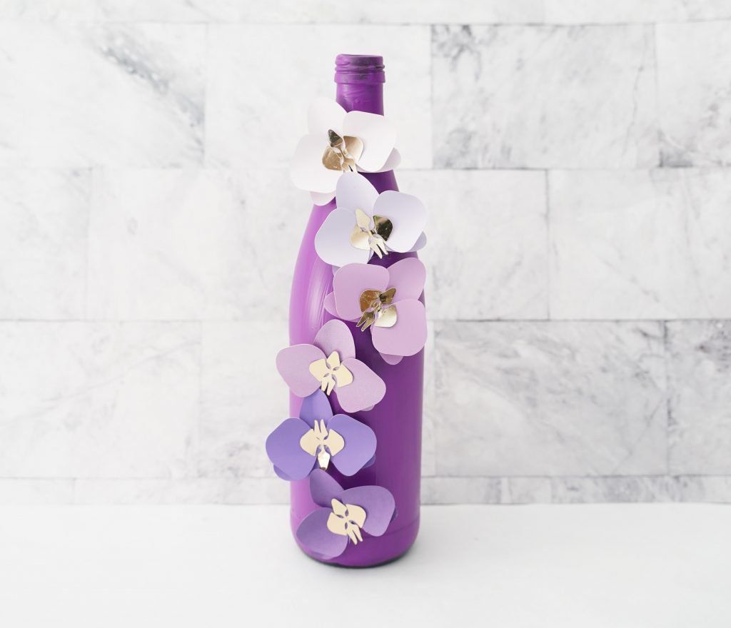 Botella pintada de morado con flores recortadas pegadas para un efecto 3D