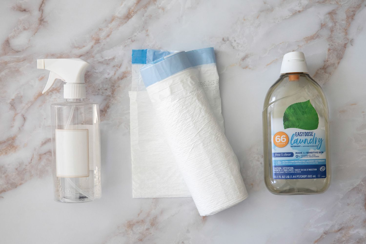Botella de spray de cristal junto a un rollo de bolsa de basura de plástico y una botella de detergente para la ropa