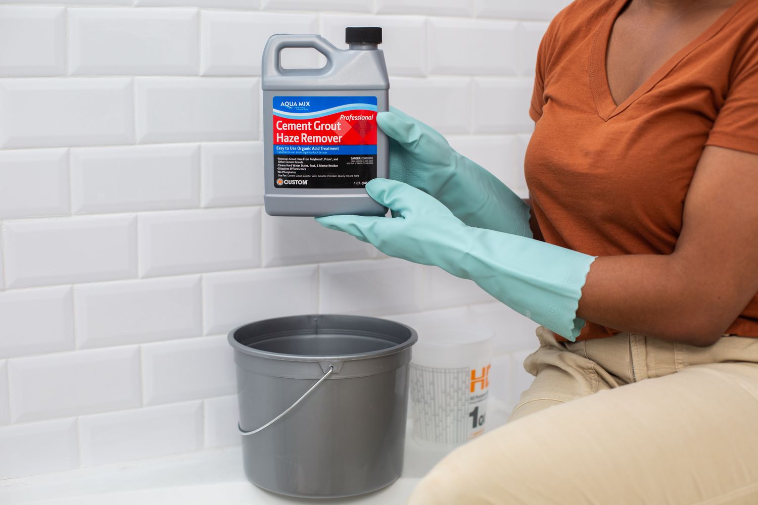 Grout haze remover bottle held over gray bucket with teal latex gloves