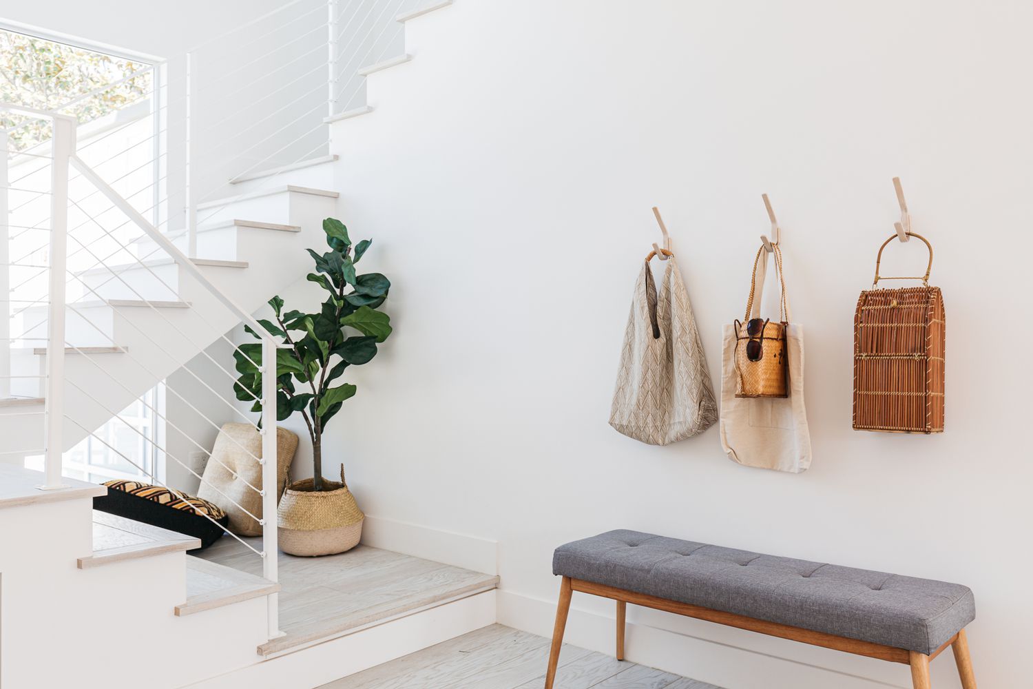 Entrada blanca con escalera cerca de la ventana con banco gris y ganchos con bolsas colgando 