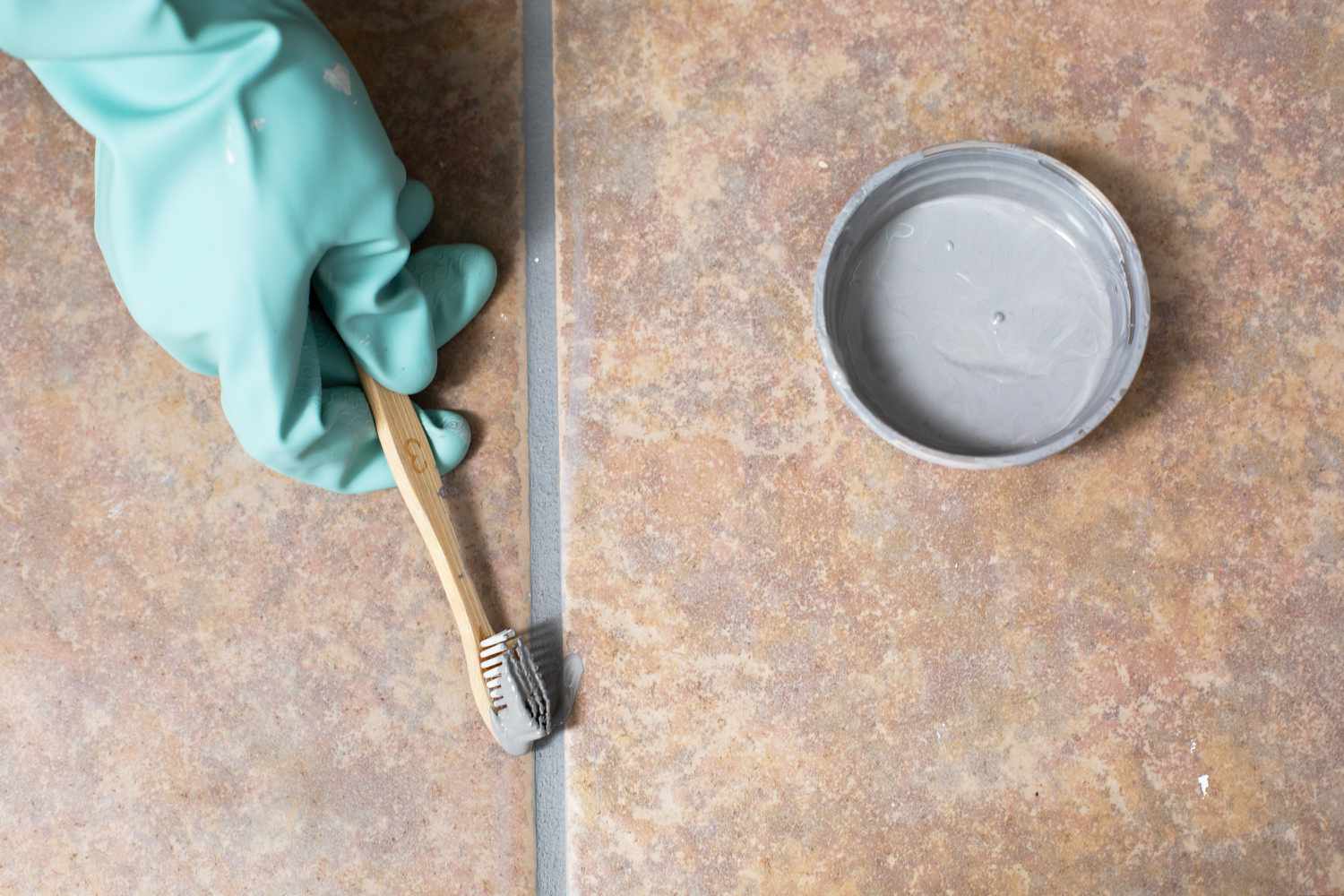 Changing grout color to gray with old toothbrush and green gloves over tiles floor