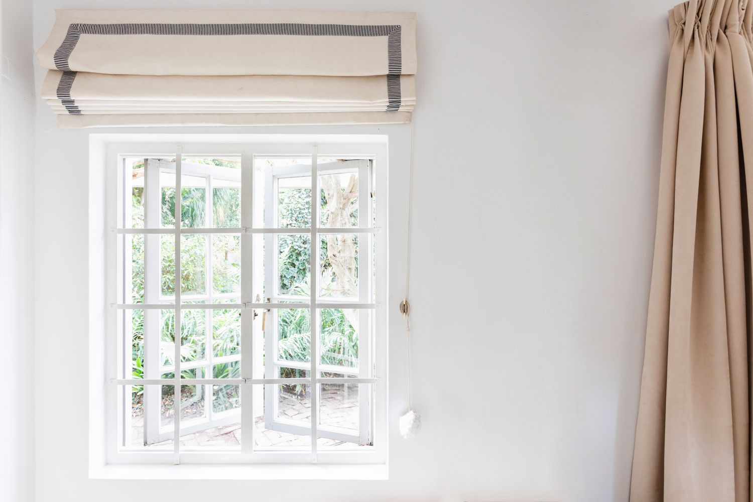 Fenster mit weißer Scheibe und gefalteten cremefarbenen und grauen Jalousien darüber und hellbrauner Gardine an der Seite