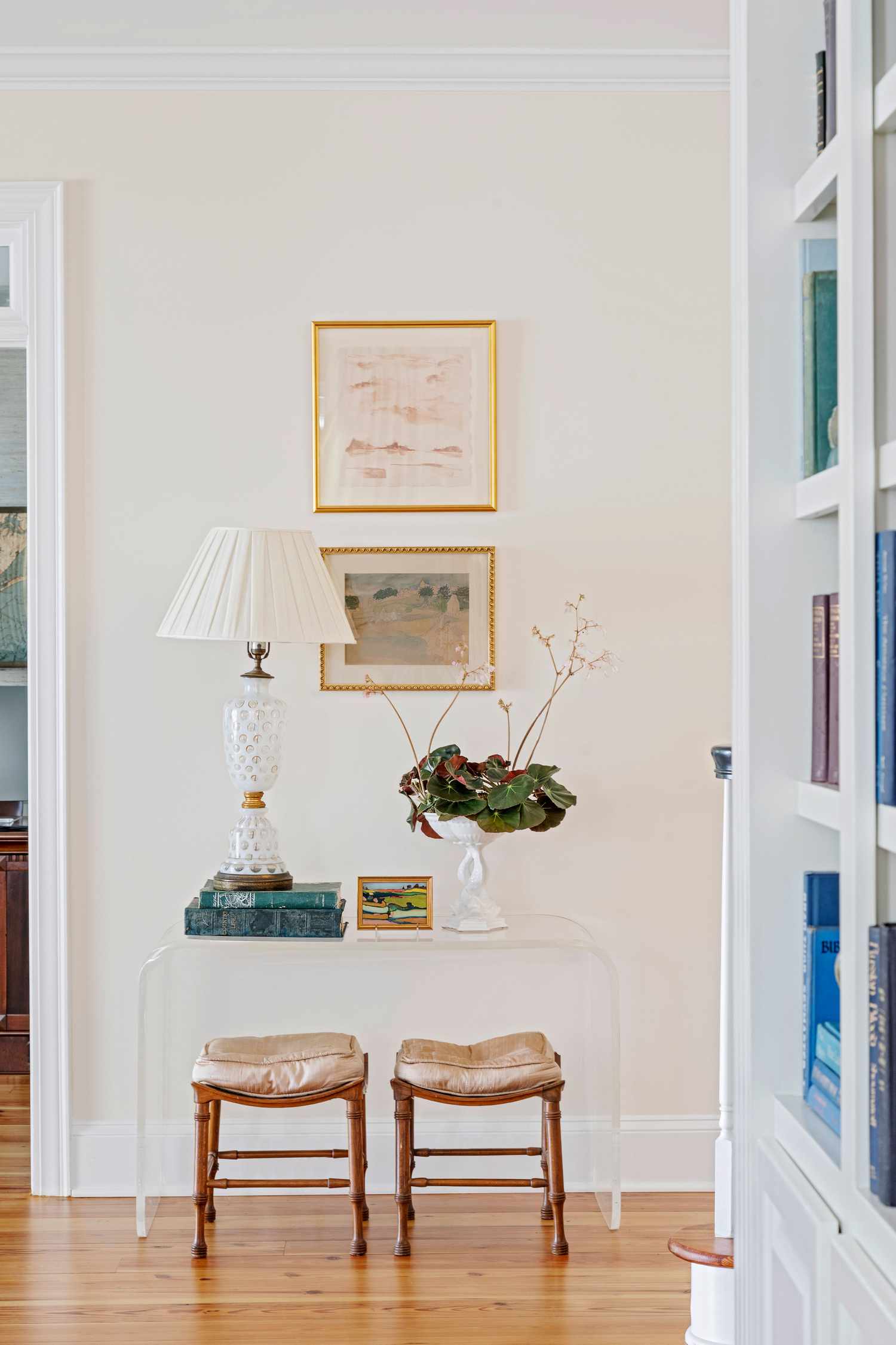 Uma mesa de console de lucite com decoração sobre ela, obras de arte acima dela e bancos abaixo