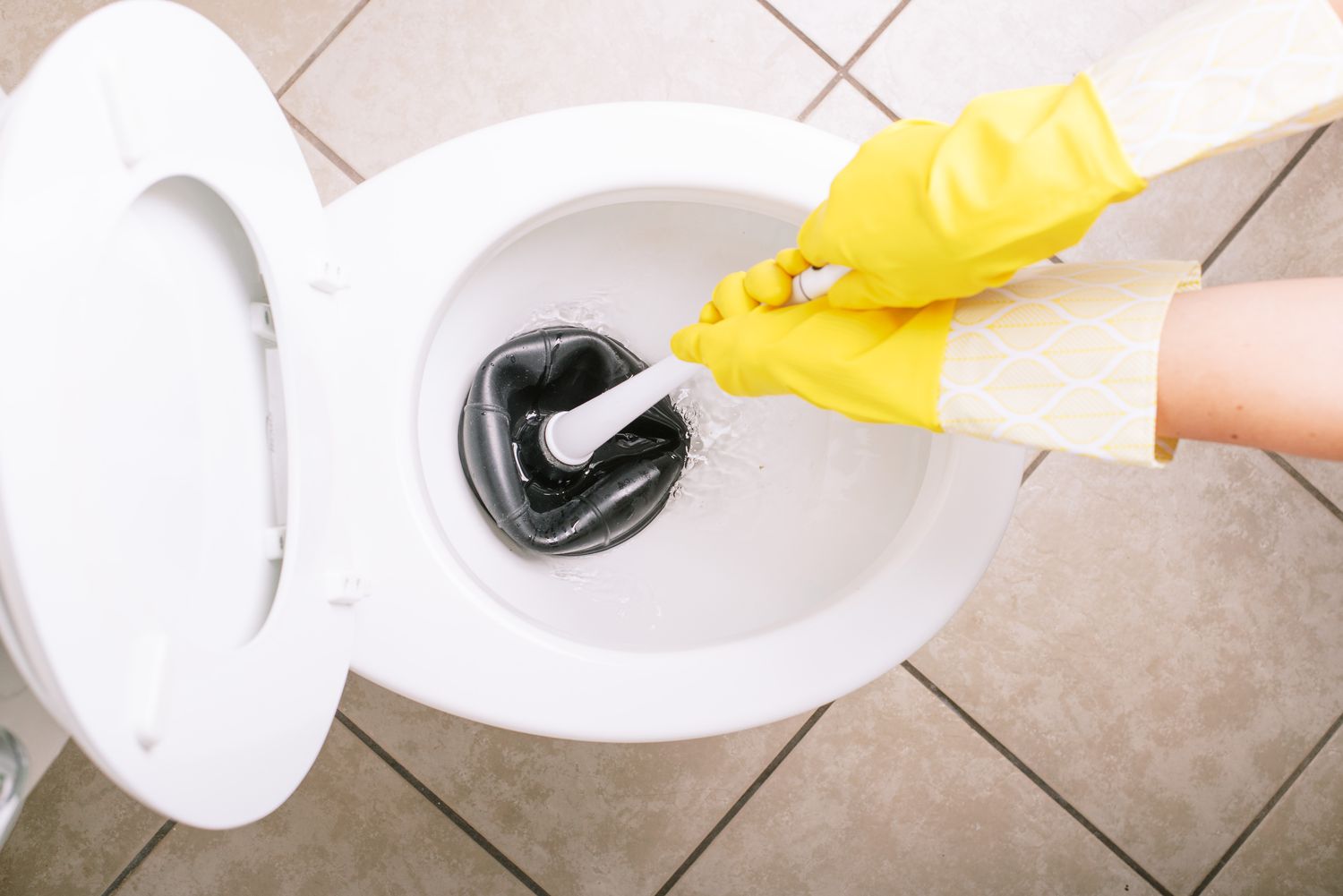 Inodoro de cerámica blanca achicando agua con desatascador negro y guantes amarillos