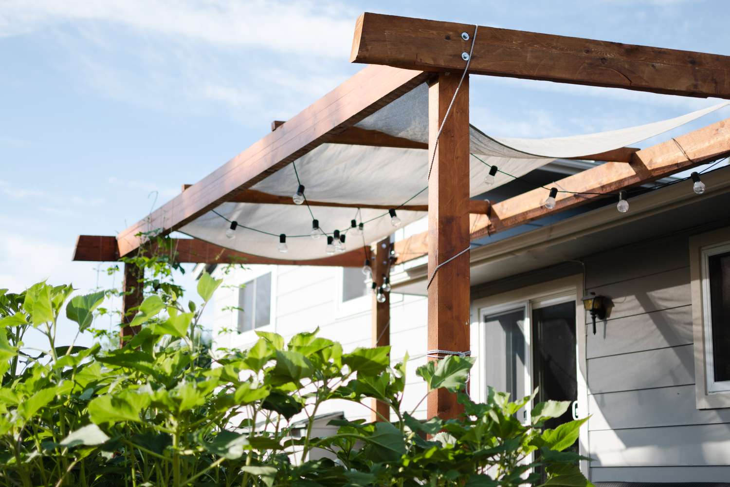 Pergola mit Holzbalken und darunter hängenden Glühbirnen mit Büschen davor