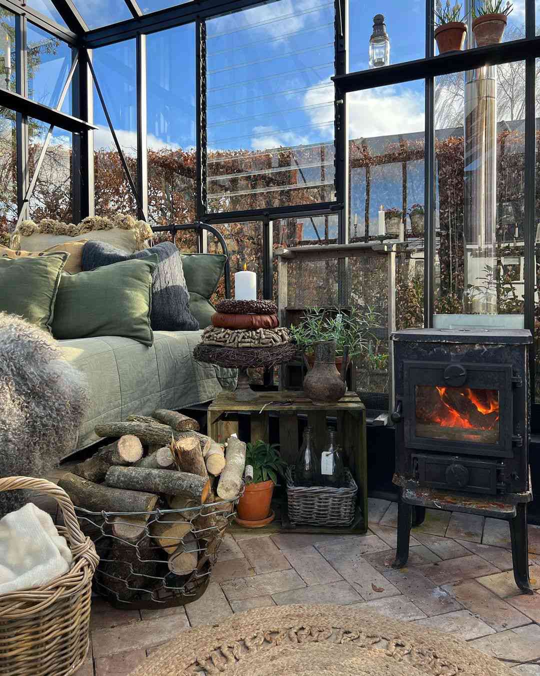 Acogedor invernadero con paredes de cristal, sofá cama y estufa de leña con cesta de leña
