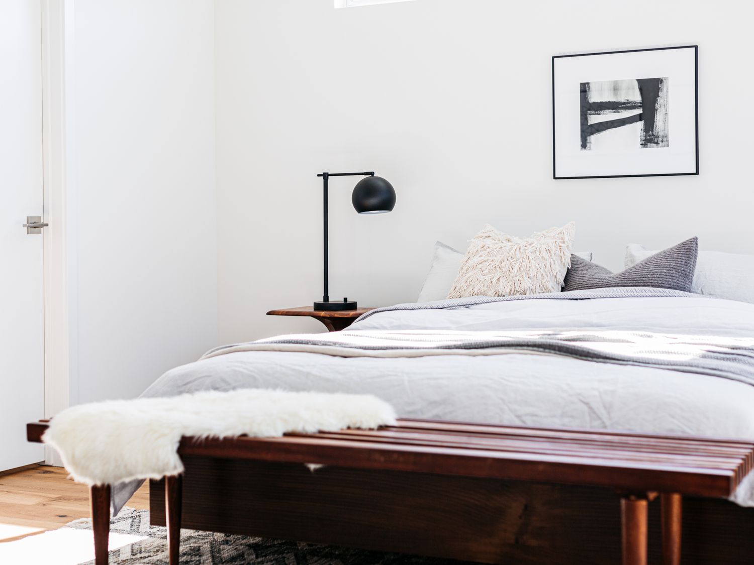 Skandinavisches Schlafzimmer mit weißen und grauen Bettlaken neben einer schwarzen Lampe mit dunkelbrauner Holzbank davor