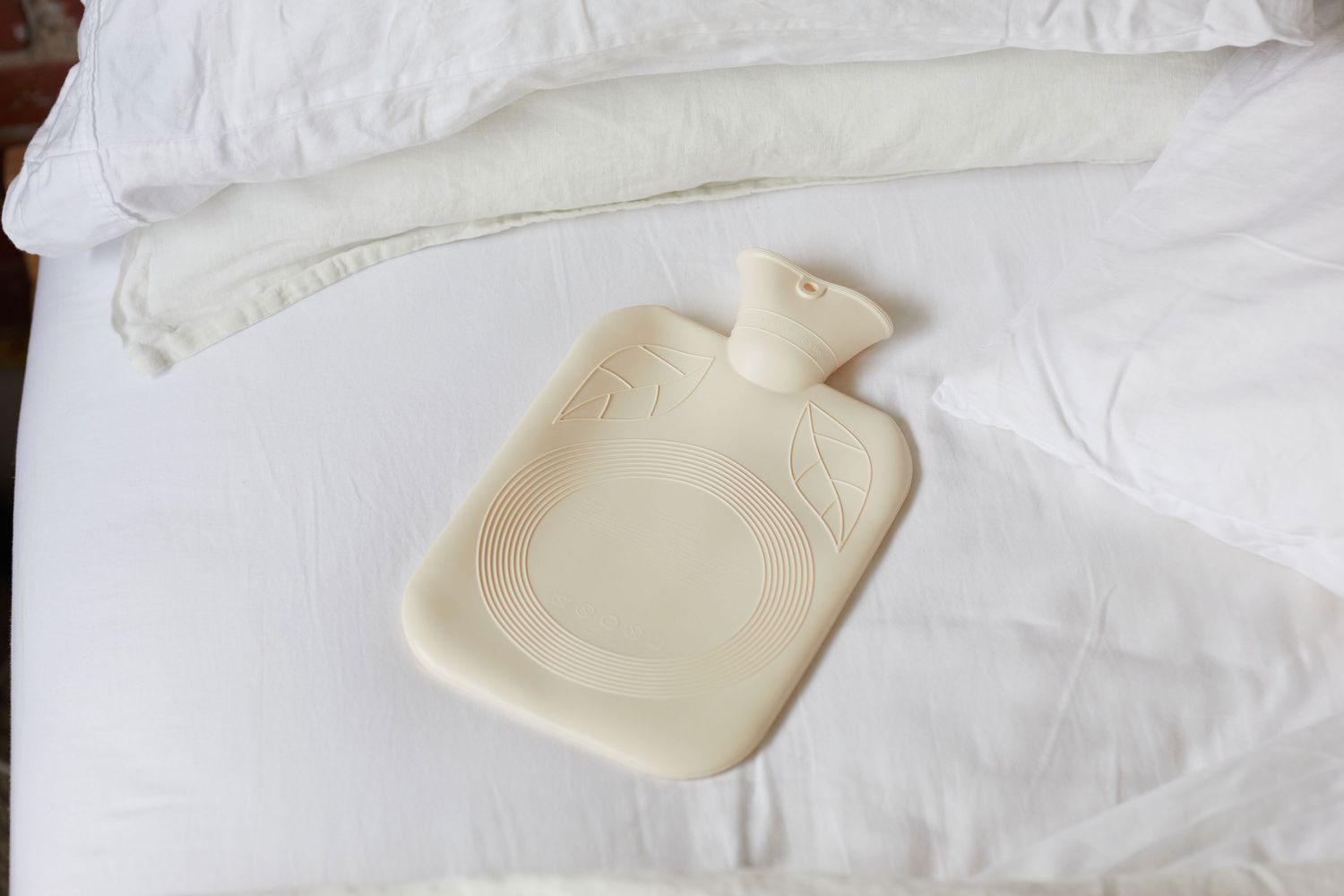 Tan-colored hot water bottle filled with cool water and placed on bed