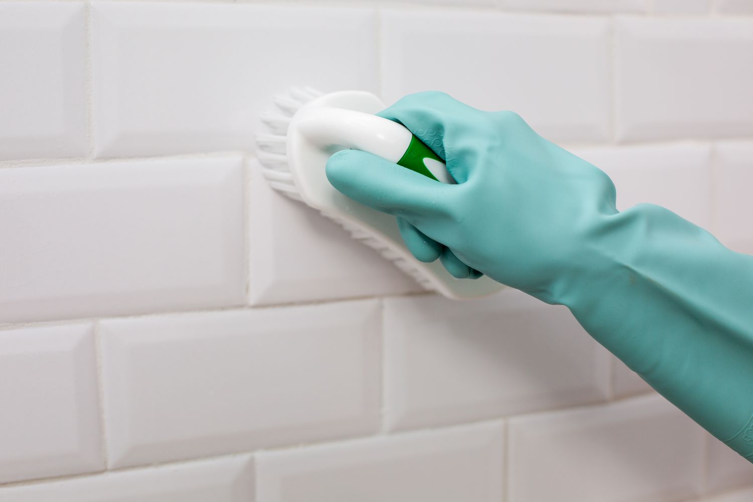 Brosse à poils de nylon frottant la solution de décapage des joints sur un mur carrelé blanc avec des gants sarcelle