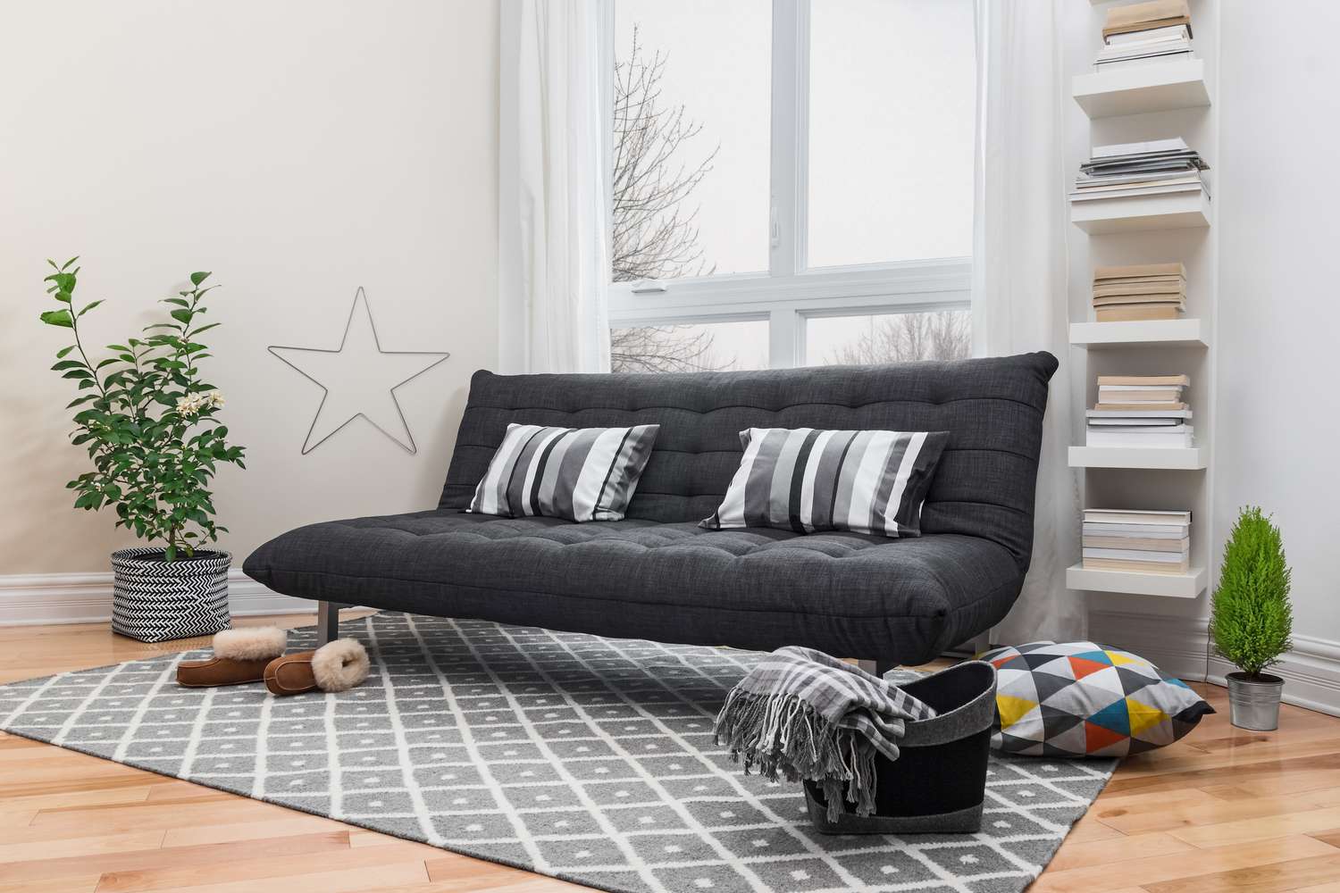 Futon cinza com almofadas listradas sobre um tapete cinza ao lado de uma estante e uma planta no chão
