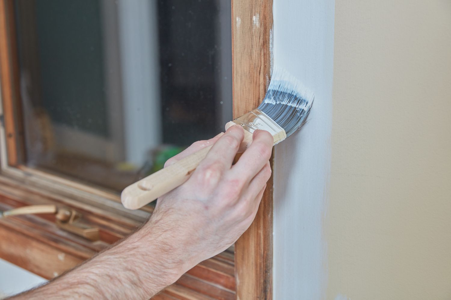 Paintbrush applying white paint to wall near wood window trim