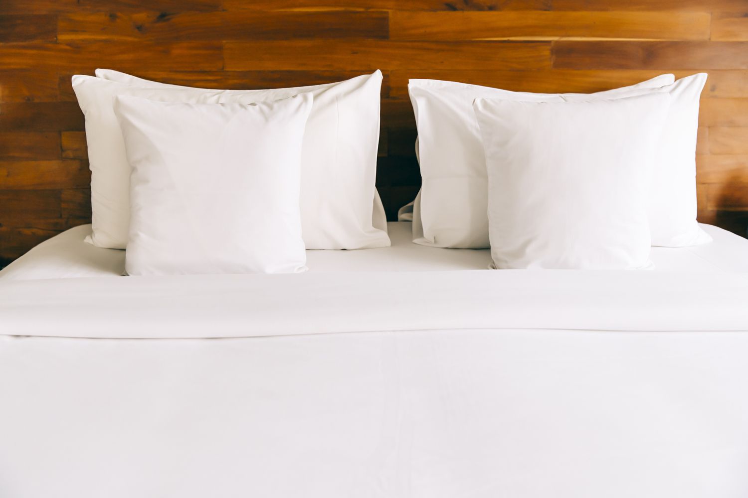 Uma cama feita com lençóis e travesseiros brancos contra uma cabeceira de madeira.