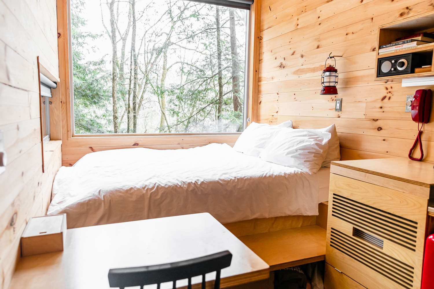 Casa minúscula de madeira com cama ao lado de uma grande janela que mostra as árvores
