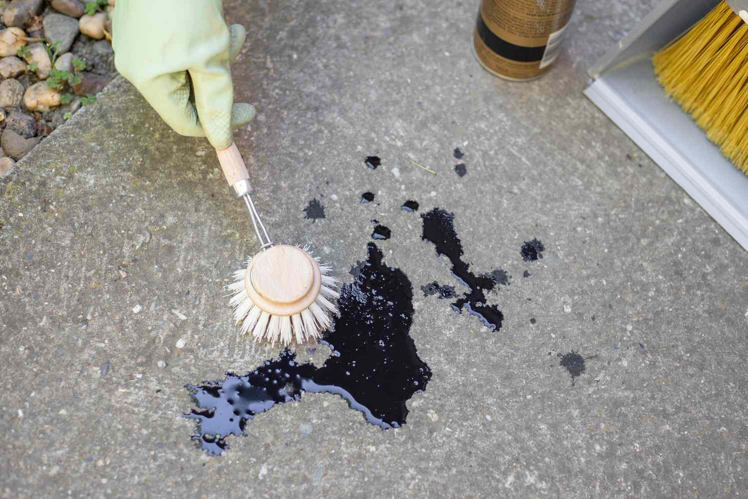Cepillo de fregar sostenido sobre una mancha de aceite negro en el suelo de hormigón con guantes