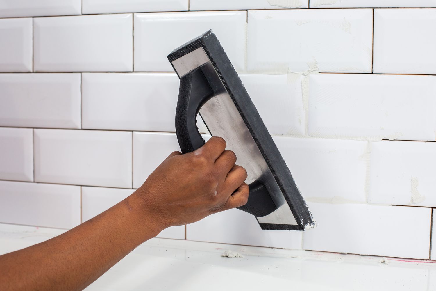 Rubber grout float applying grout to white ceramic wall tiles