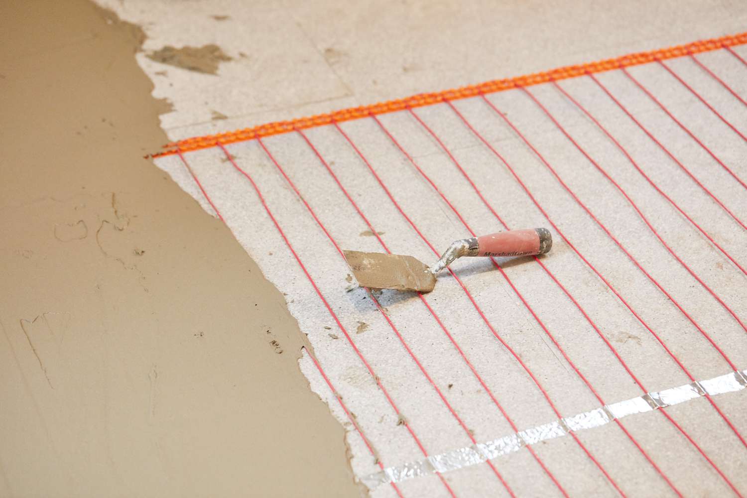 Système de plancher chauffant électrique avec fils orange recouverts partiellement de ciment-colle et de grattoir