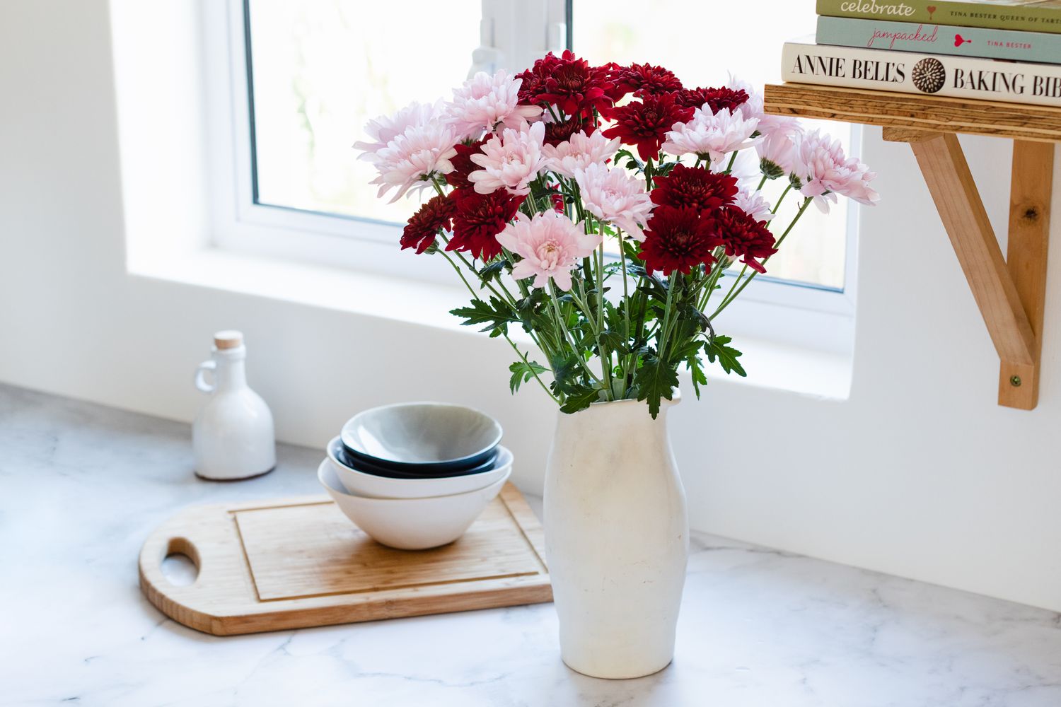 Rosa und rote Chrysanthemenblüten in einer hohen weißen Vase neben Fenster und Bücherregal