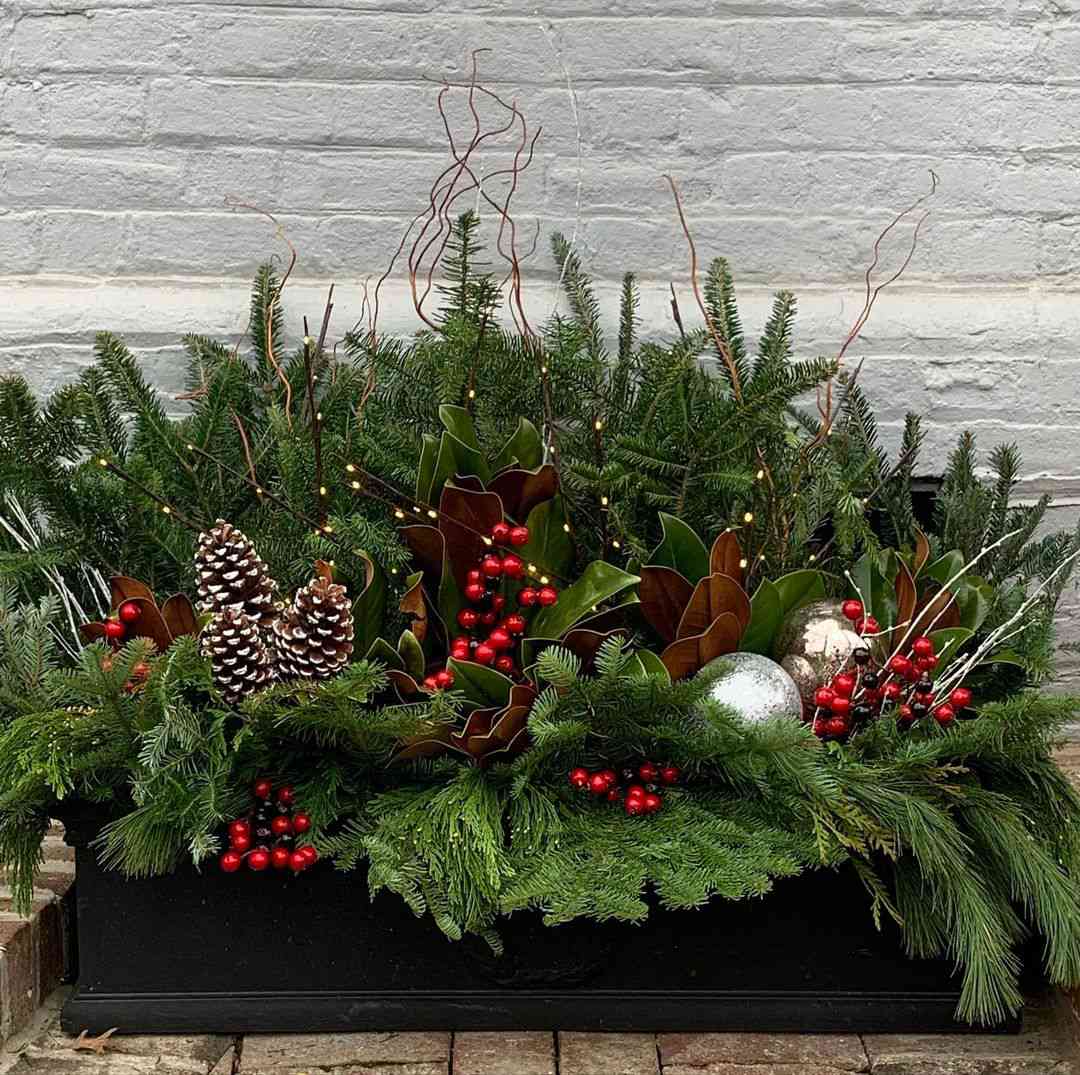 jardinera de invierno con hojas perennes, bayas rojas, piñas tocadas de blanco y adornos redondos de cristal plateado