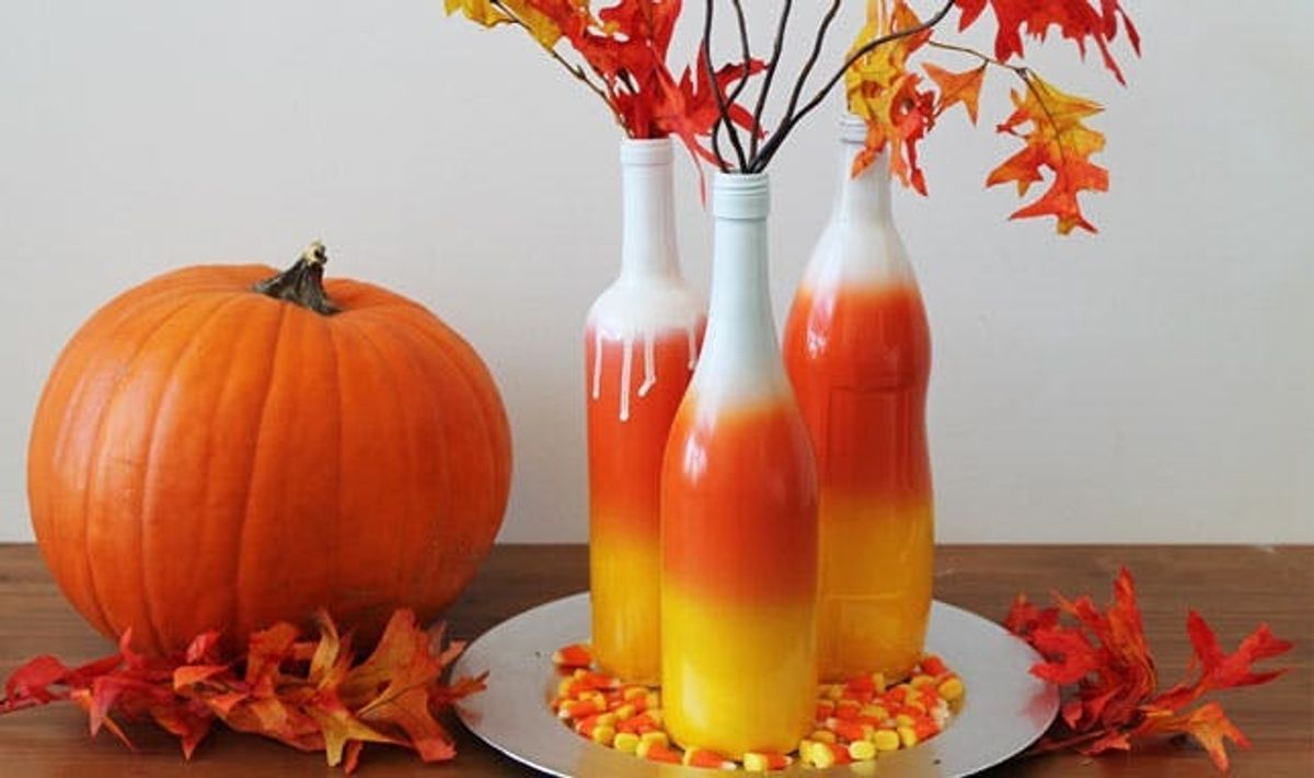 Tres botellas pintadas con spray para que parezcan maíz de caramelo con decoración otoñal a su alrededor