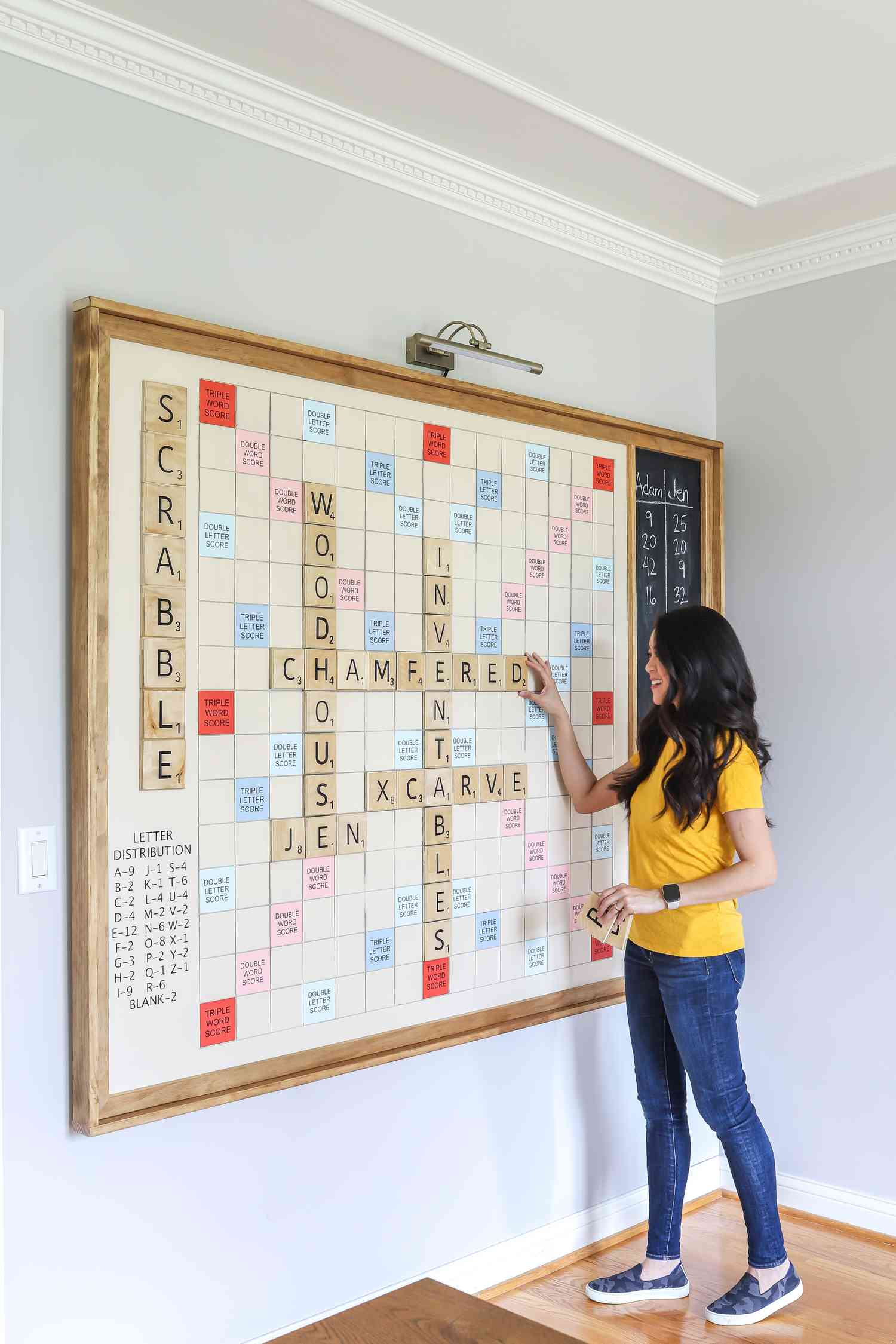 Jen Woodhouse posa ao lado de seu tabuleiro de Scrabble gigante DIY montado em uma parede
