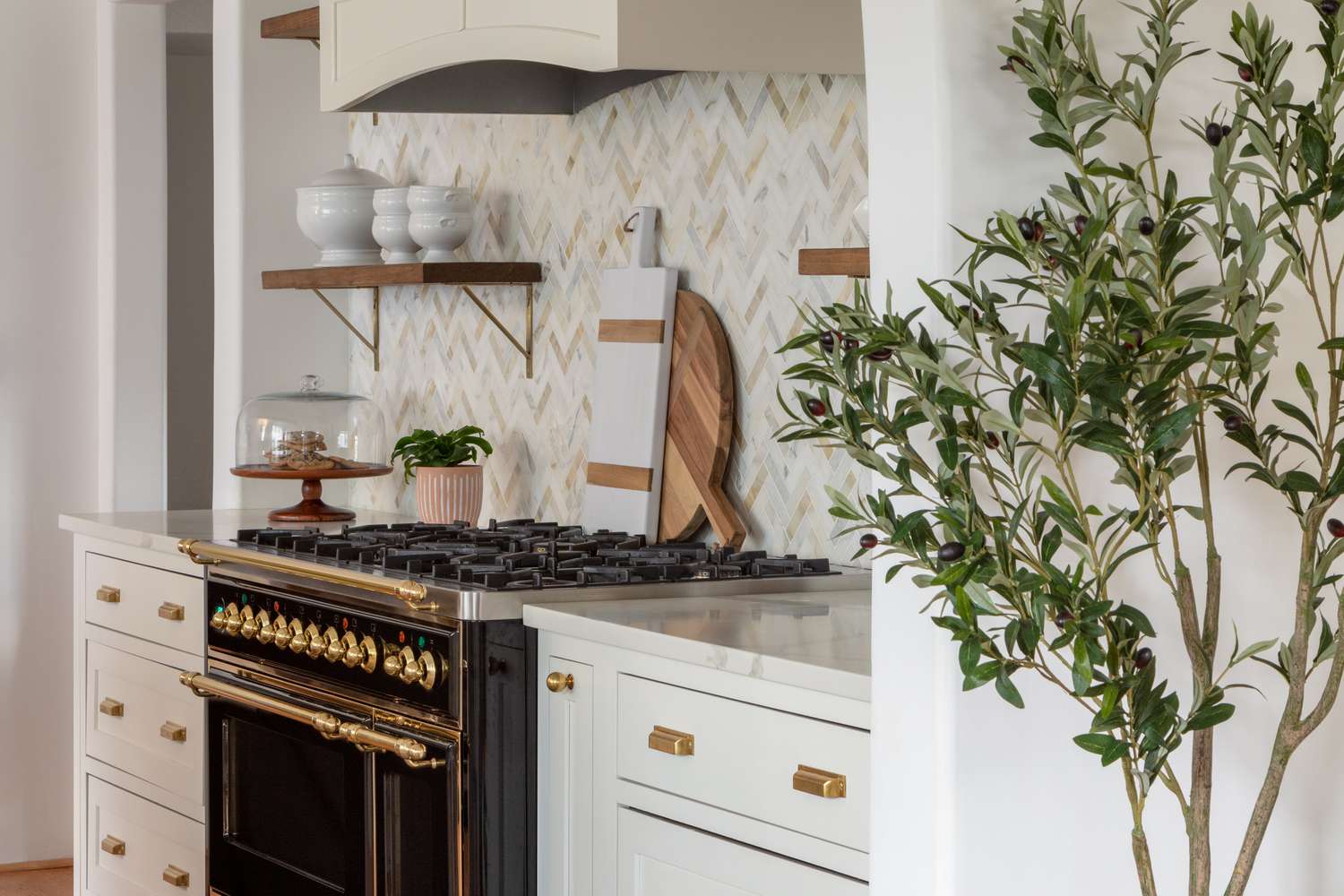 Die Küche im Haus von Erin Coren verfügt über einen schwarzen Herd, offene Regale und weiße Schränke mit goldenen Beschlägen, die wie Zehentreter aussehen