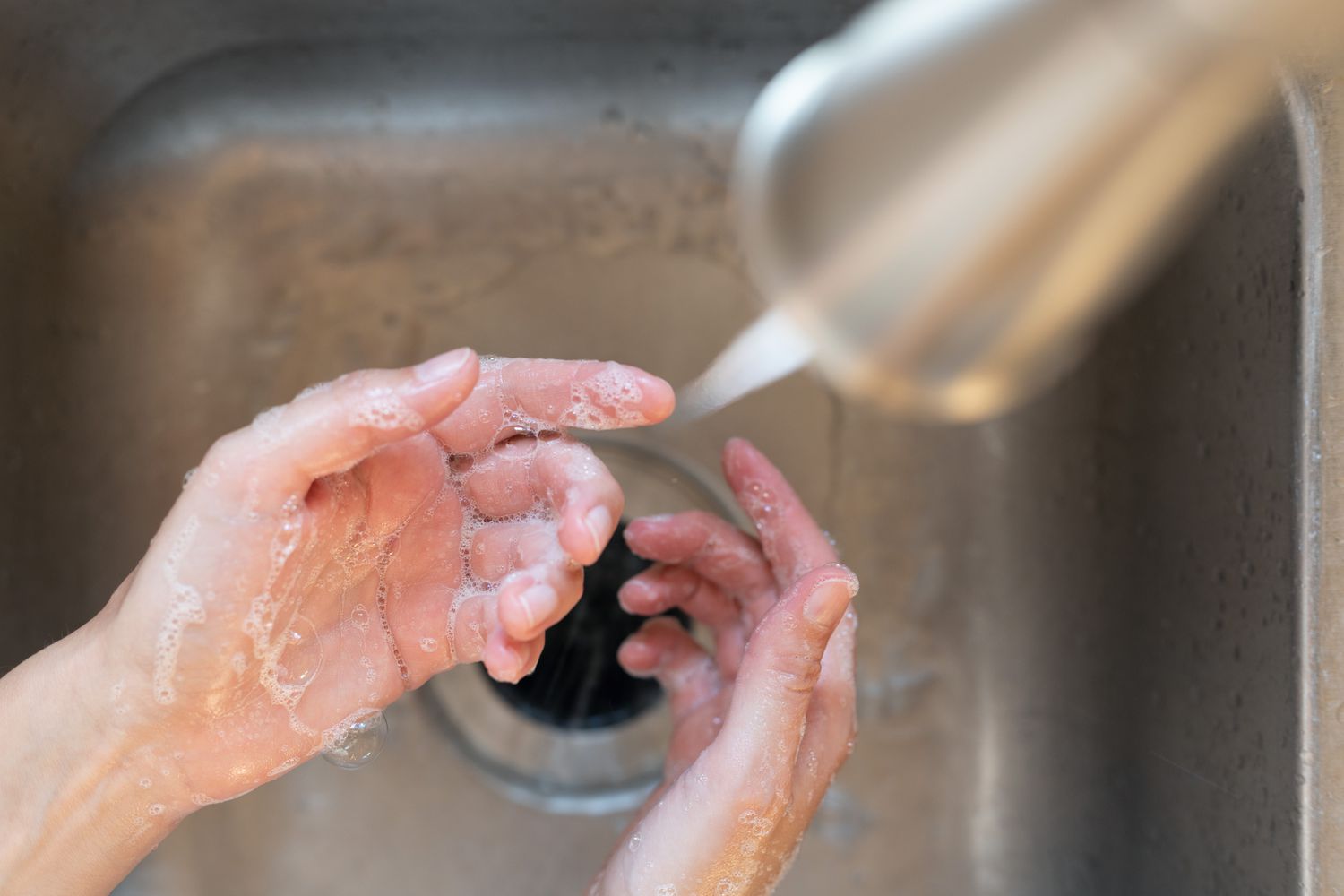 Hands covered in warm soapy water in sink and below robinet