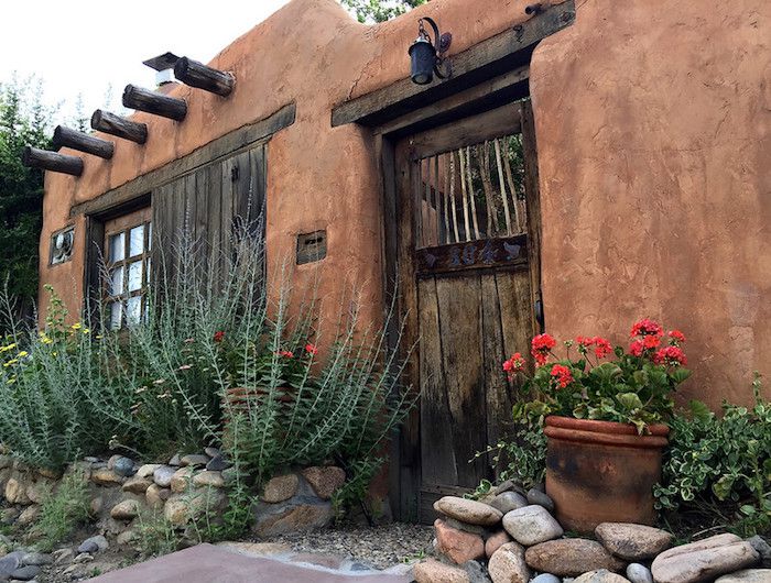 Maison en adobe avec porte en bois, sauge russe poussant sur le mur rocheux près de l'entrée et pot de géraniums rouges.