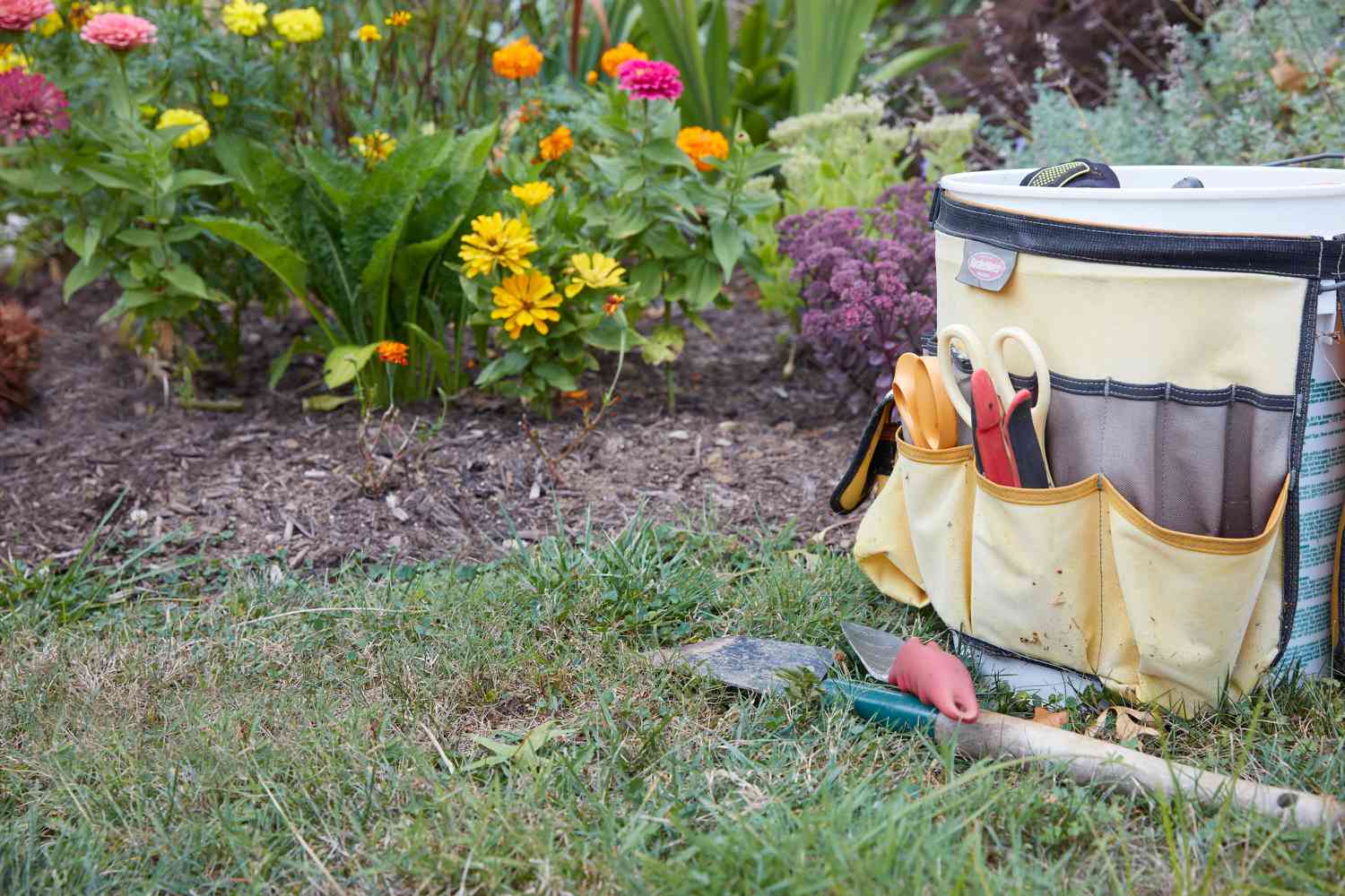 Bolsa de jardinagem com ferramentas nos bolsos e pás na frente, ao lado do jardim de flores