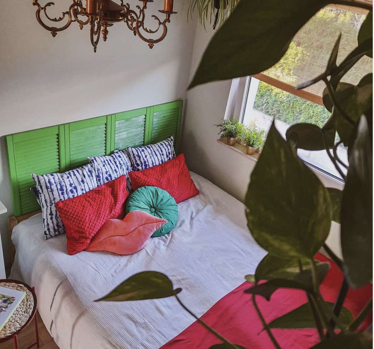 dormitorio con cabecero verde brillante y plantas, almohadas y manta rojas, elegante araña de bronce