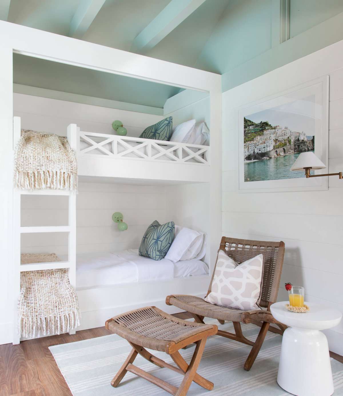 quarto com beliches, teto azul-petróleo com vigas de madeira expostas, paredes brancas, marrom e cadeira de vime com banquinho para os pés