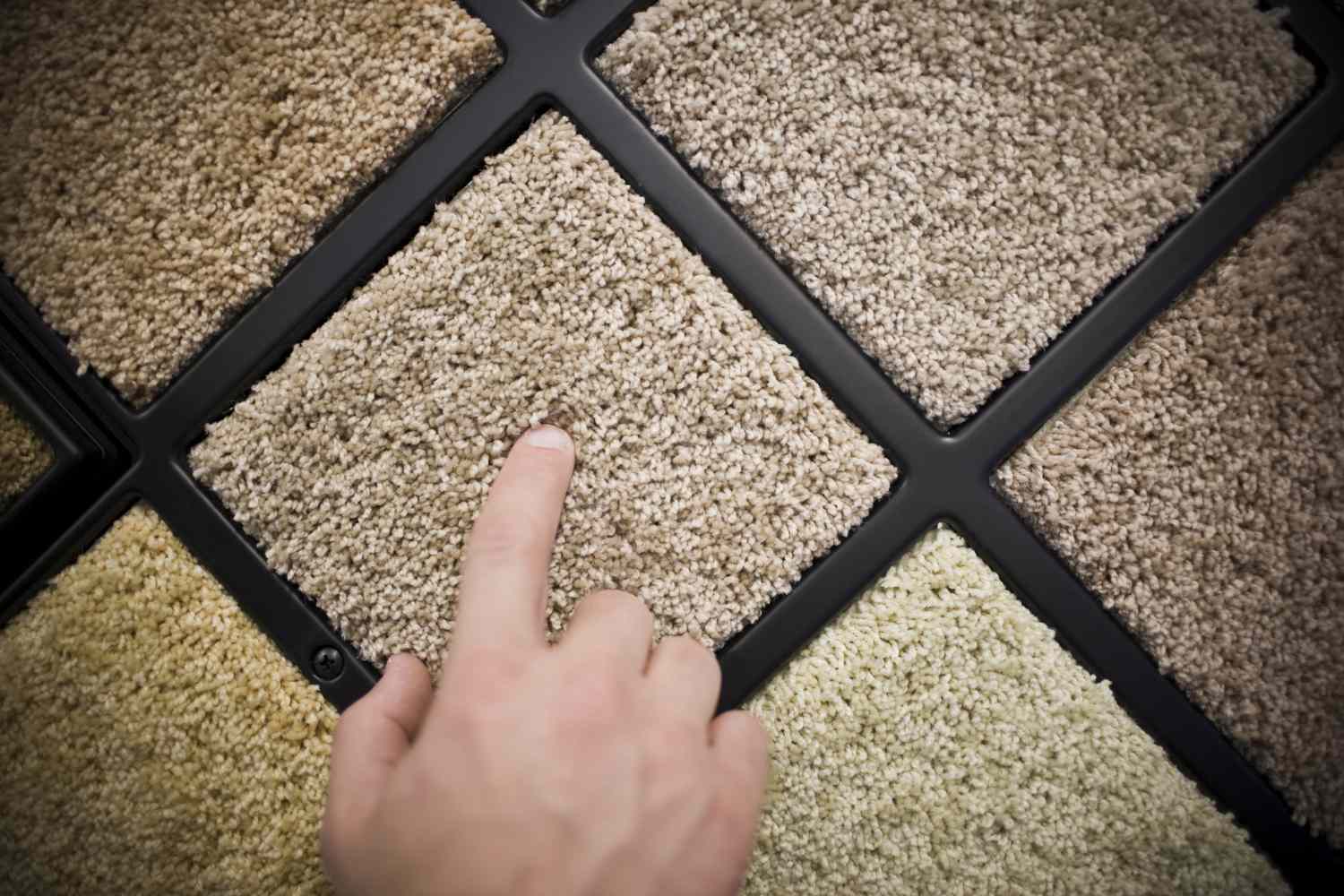 Finger pointing at one of several beige carpet samples on a carpet sample wall