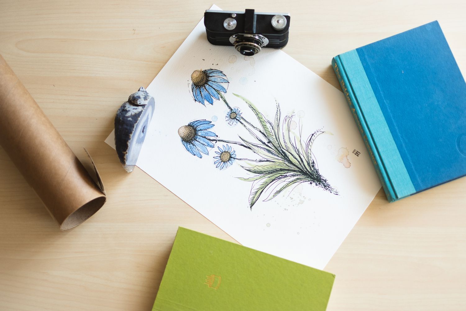 Plakat mit blauen Blumen auf einer Fläche mit Kamera, Büchern und Felsen zum Abflachen ausgelegt 