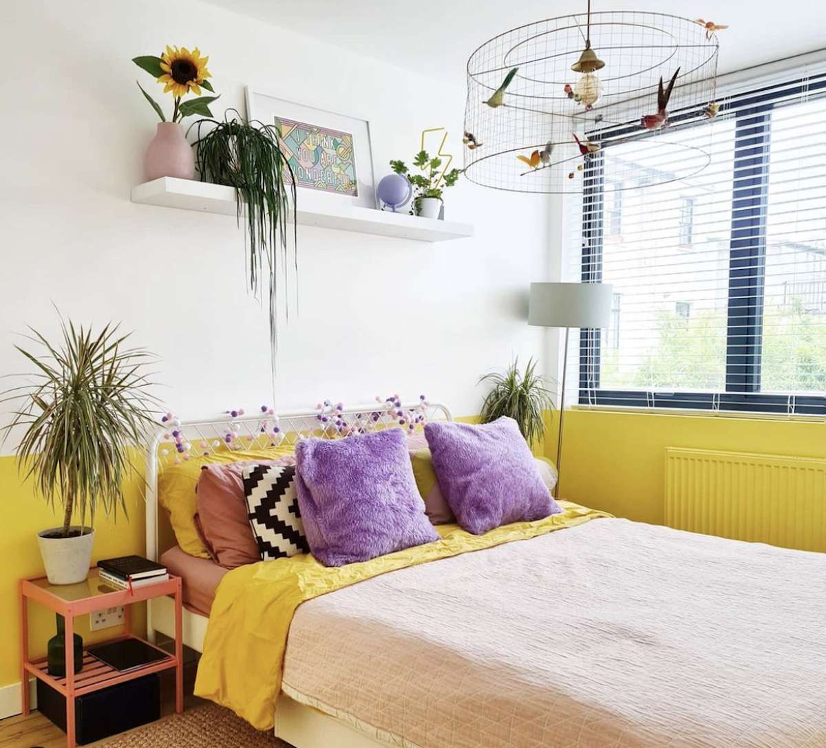 dormitorio morado y amarillo con paredes blancas, estantería flotante con plantas y obras de arte, lámpara de araña que parece una jaula de pájaros
