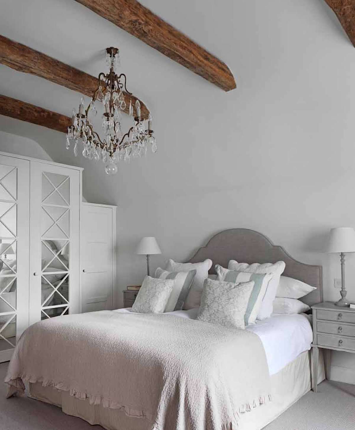 quarto com paredes brancas, cores neutras com leves toques de azul, vigas de madeira expostas no teto, lustre ornamentado, estilo toscano