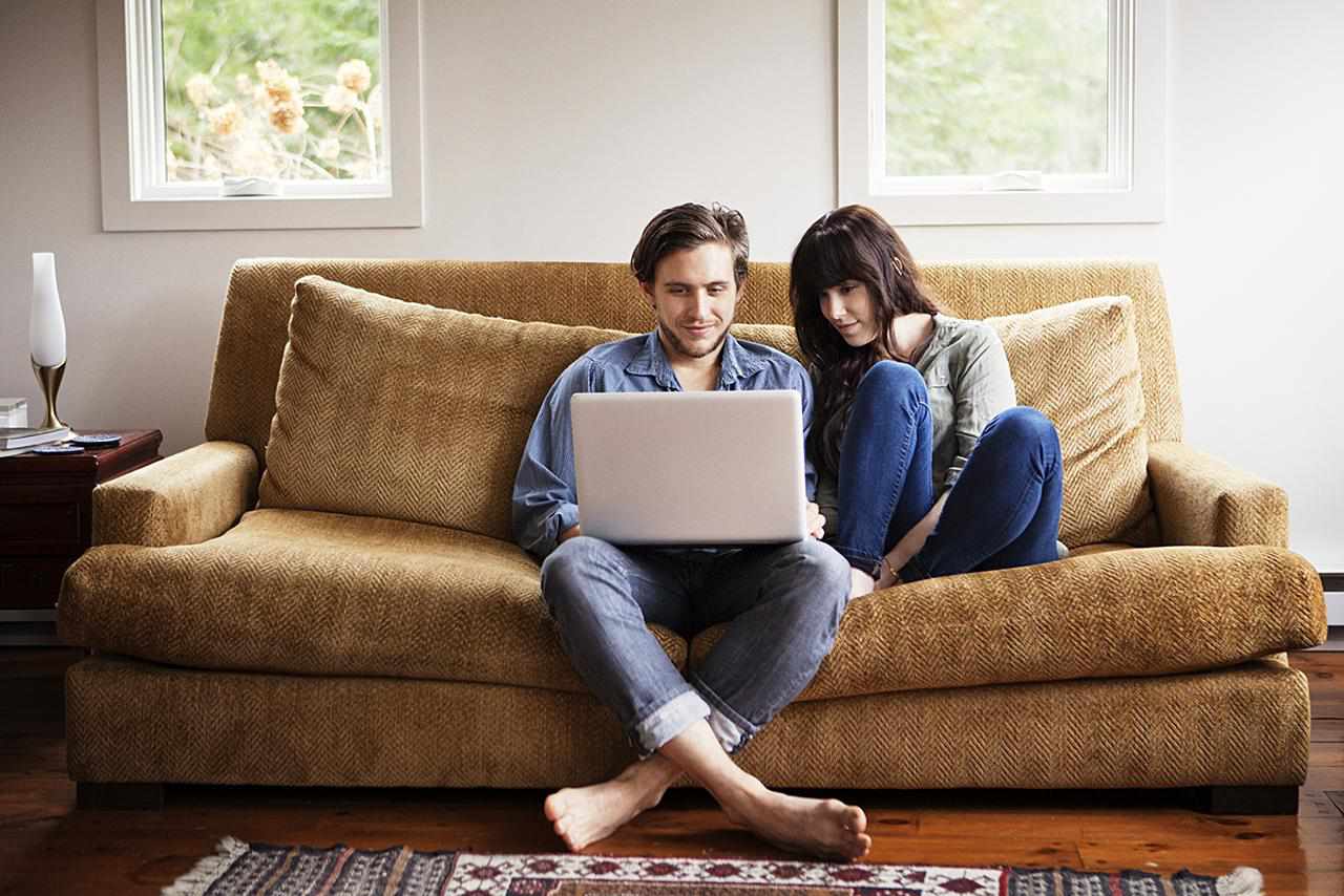 Jovem casal sentado em um sofá enquanto o rapaz está em um laptop.