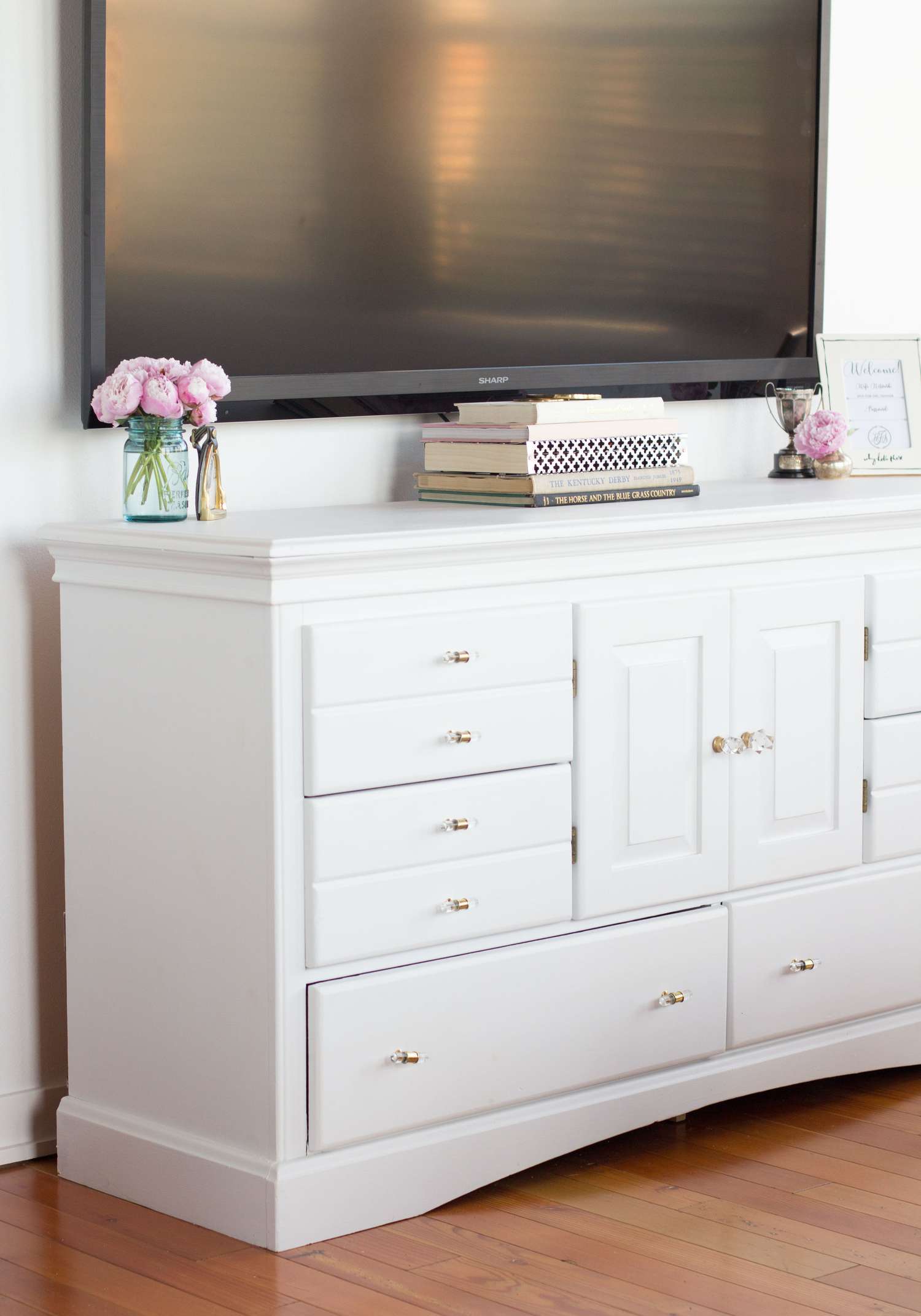 Gran televisor sobre un mueble consola blanco con libros y jarrones de flores