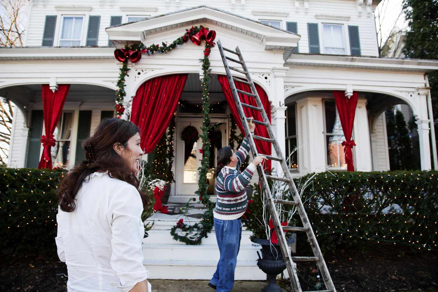 Pareja colgando guirnaldas en una casa con una escalera por Navidad.