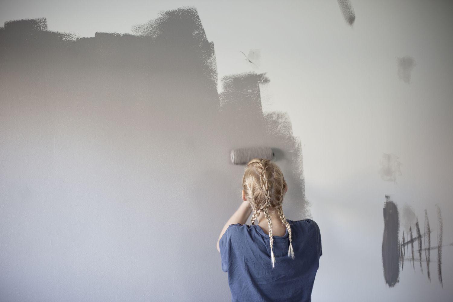 Blonde Frau mit Zöpfen streicht eine weiße Wand mit einem Farbroller grau