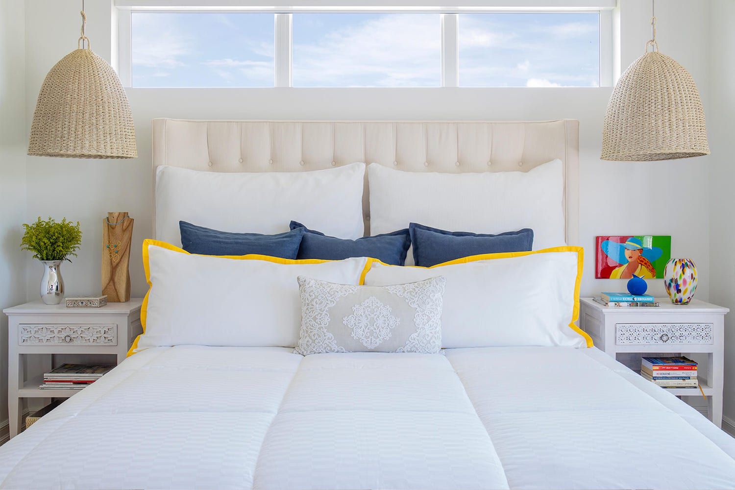 überwiegend weißes, neutrales Schlafzimmer mit Rattan-Hängelampen, kleinen Farbtupfern, offenen Fenstern über dem Bett